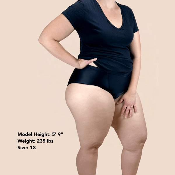 A model wearing a black short-sleeve V-neck top and and Hem Support Wear's High Waisted Brief prolapse support underwear in black. Text in the lower left corner reads 'Model Height: 5' 9" Weight: 235 lbs Size: 1X'.
